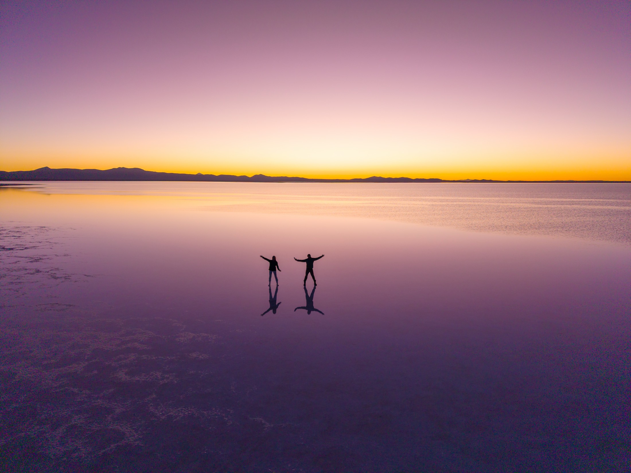 Two people walk together across a mirror-like salt flat at sunset